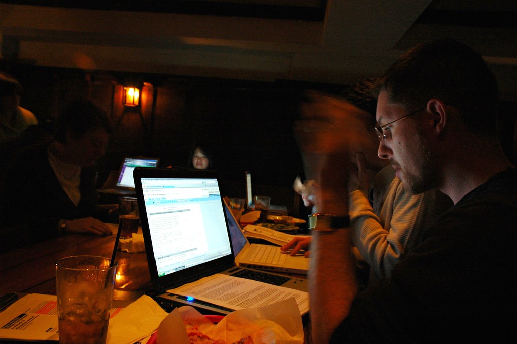 A student typing a paper on a laptop under a desk lamp in a dorm room late at night